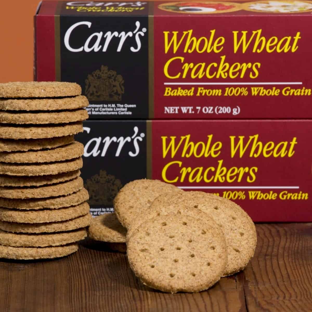 Carr's whole wheat crackers stacked on table in front of two packages of crackers.