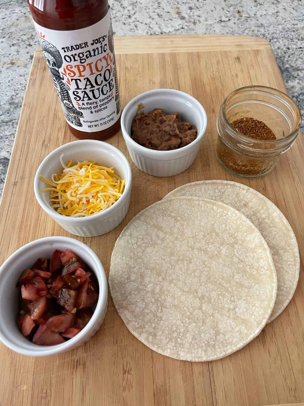 Ingredients including taco sauce, shredded cheese, refried beans, taco seasoning, chopped tomatoes and two corn tortillas on bamboo cutting board.