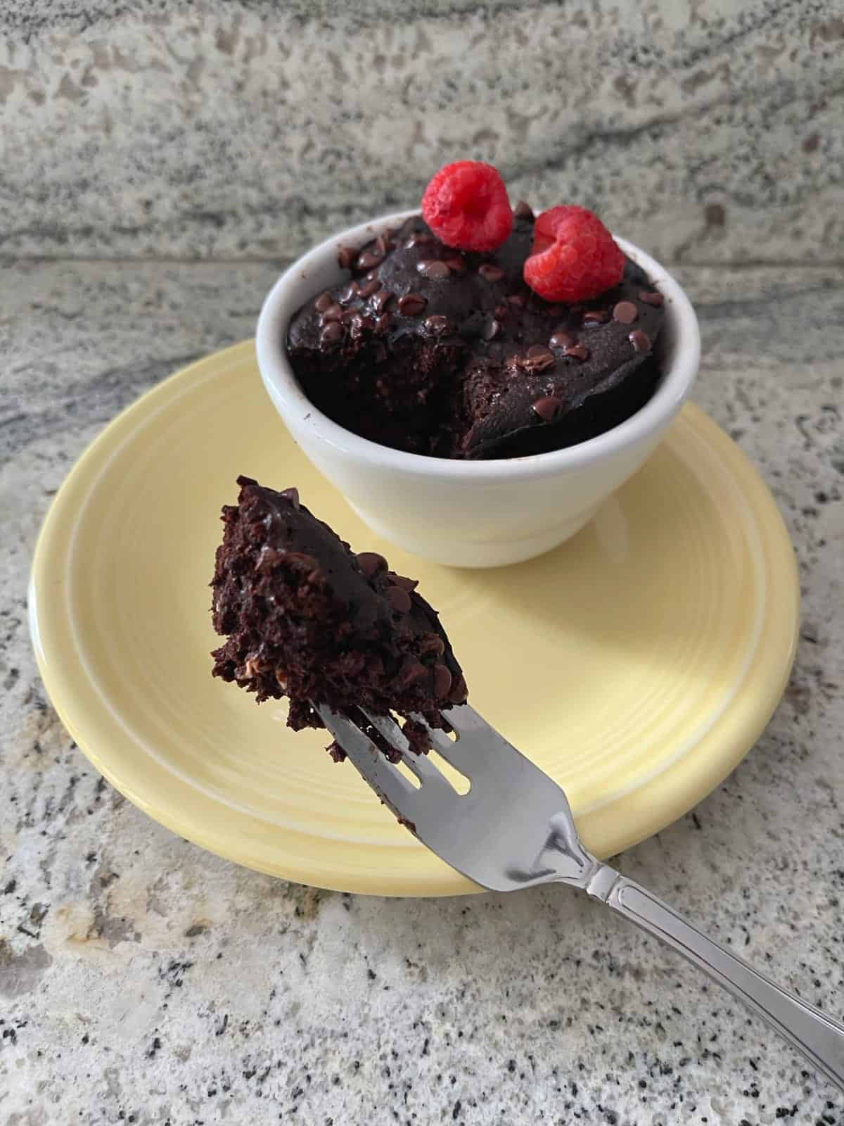 Microwave mocha mug cake topped with fresh raspberries on yellow plate with fork full of cake.