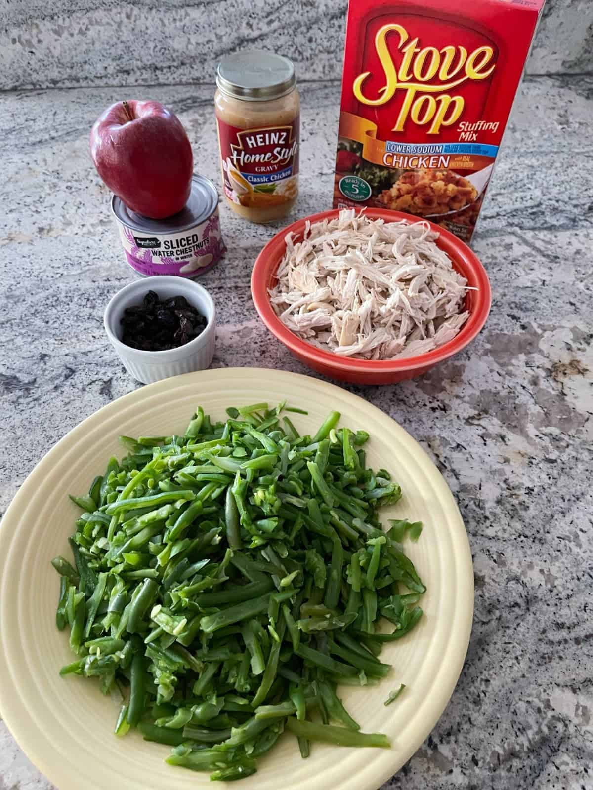 Stove Top chicken stuffing mix, shredded chicken breast, french-style green beans, raisin, apple and chicken gravy on granite counter.