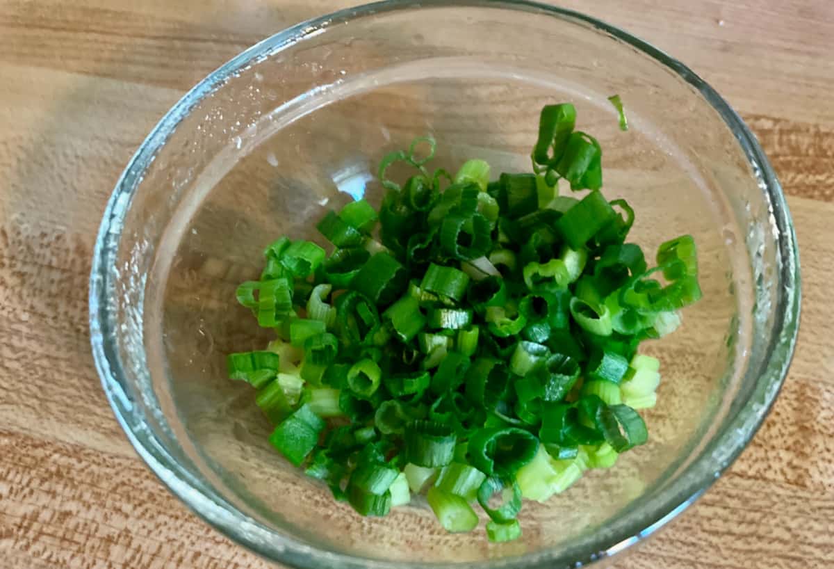 Small glass bowl with sliced green onions on butcher block counter.
