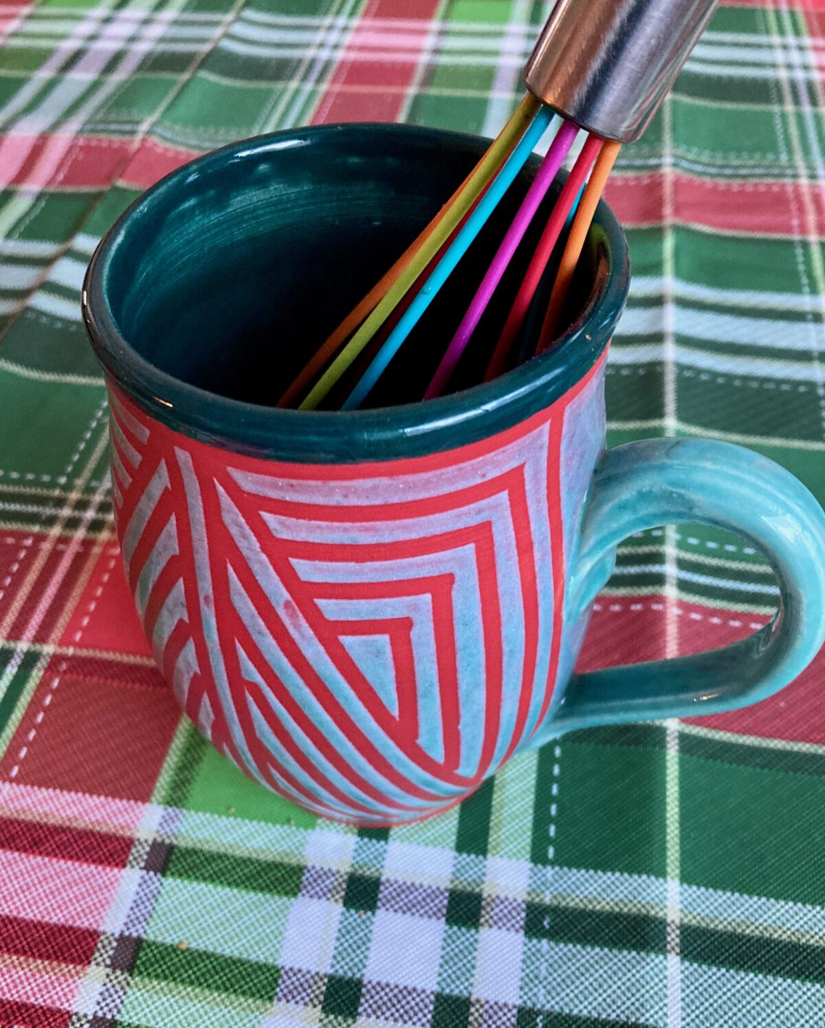 Red and green mug with wire whisk on holiday plaid tablecloth.