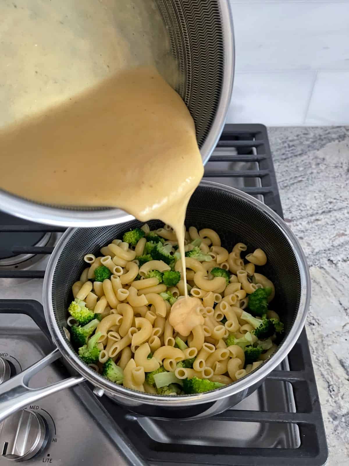 Pouring cheese sauce over cooked macaroni and broccoli in saucepan.