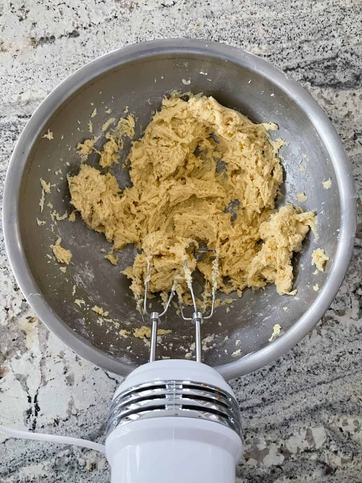 Beating coconut macaroon kiss cookie dough with electric mixer in stainless mixing bowl.