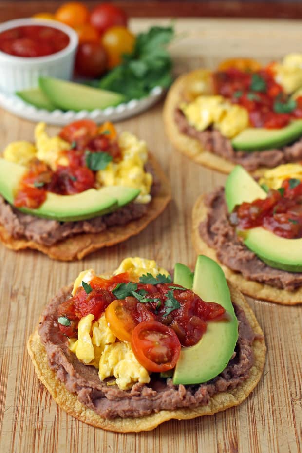 Corn tostadas topped with refried beans, scrambled eggs, avocado slices, sliced grape tomatoes, salsa and cilantro on wooden cutting board.