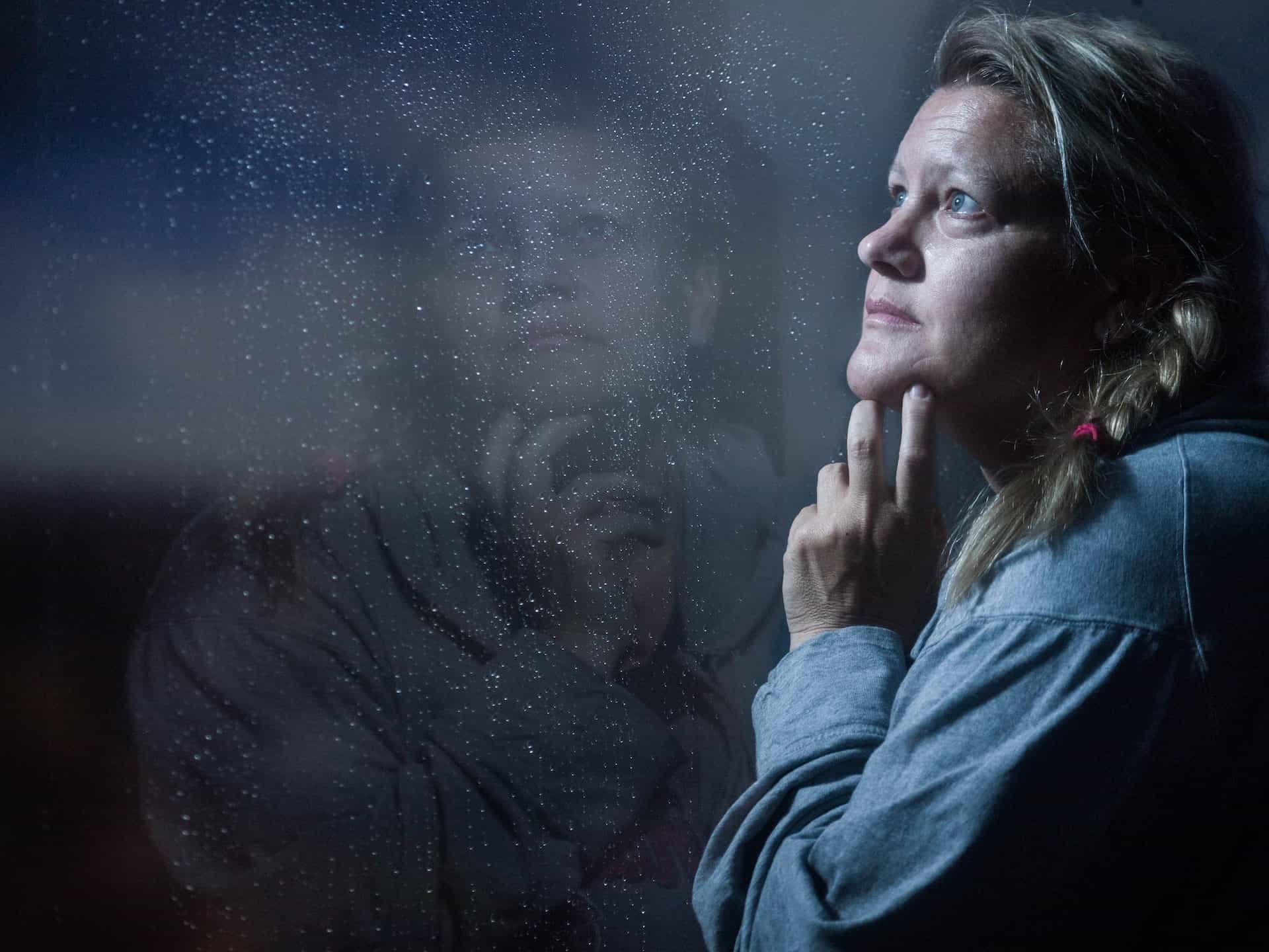 Contemplative woman in blue shirt looking out window with reflection