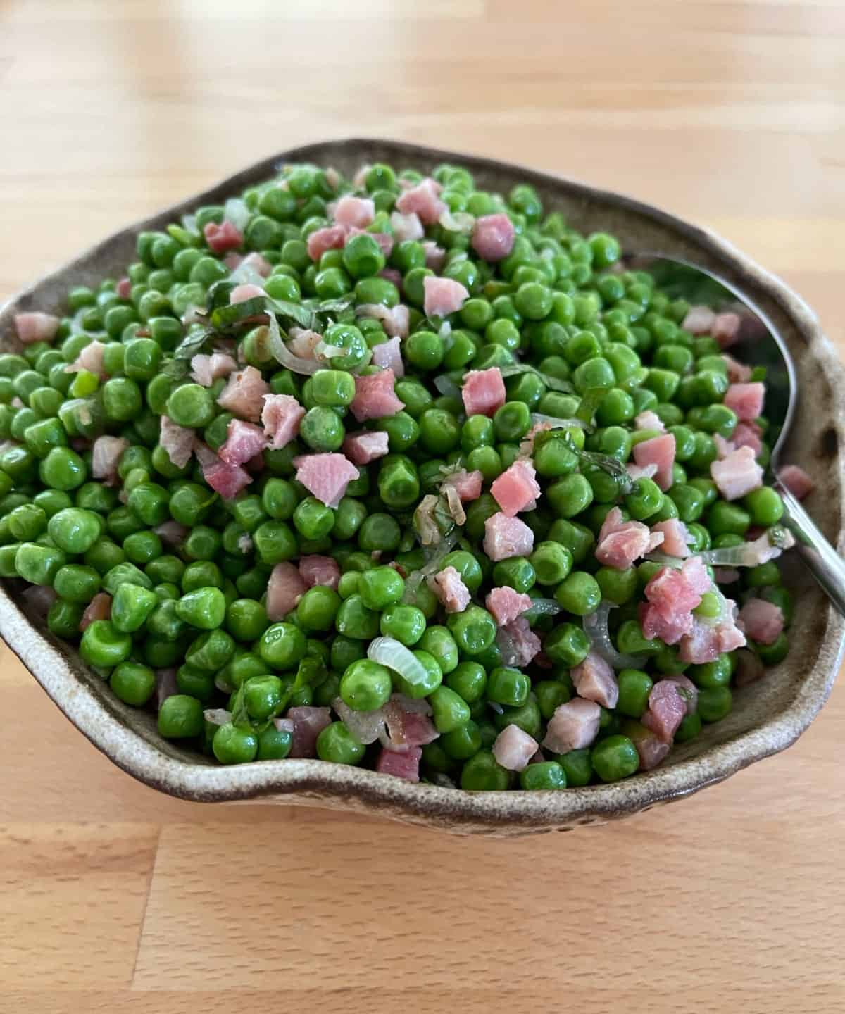 Fresh cooked peas with pancetta in brown ceramic bowl with serving spoon.