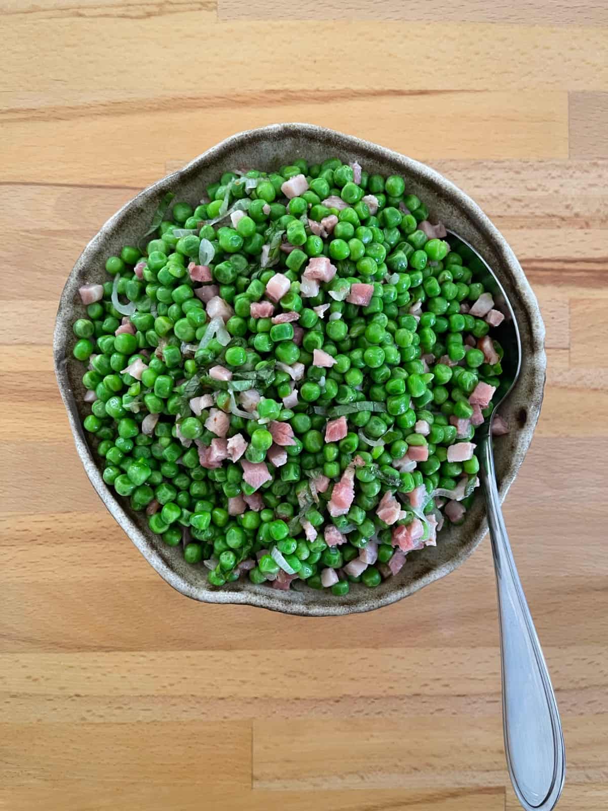 Peas with pancetta and fresh mint in ceramic bowl with serving spoon.
