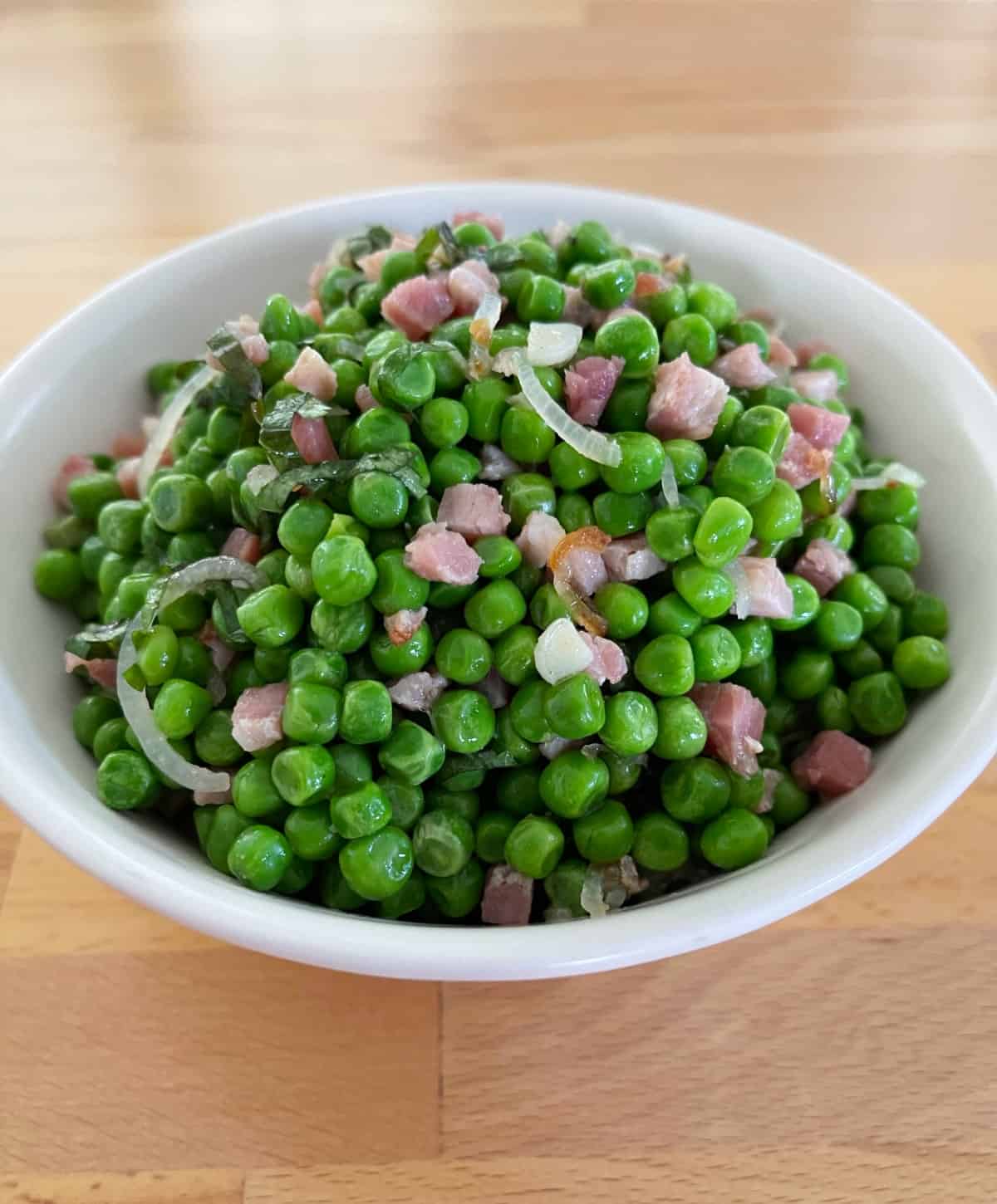 Peas and pancetta in white serving bowl on wood table.