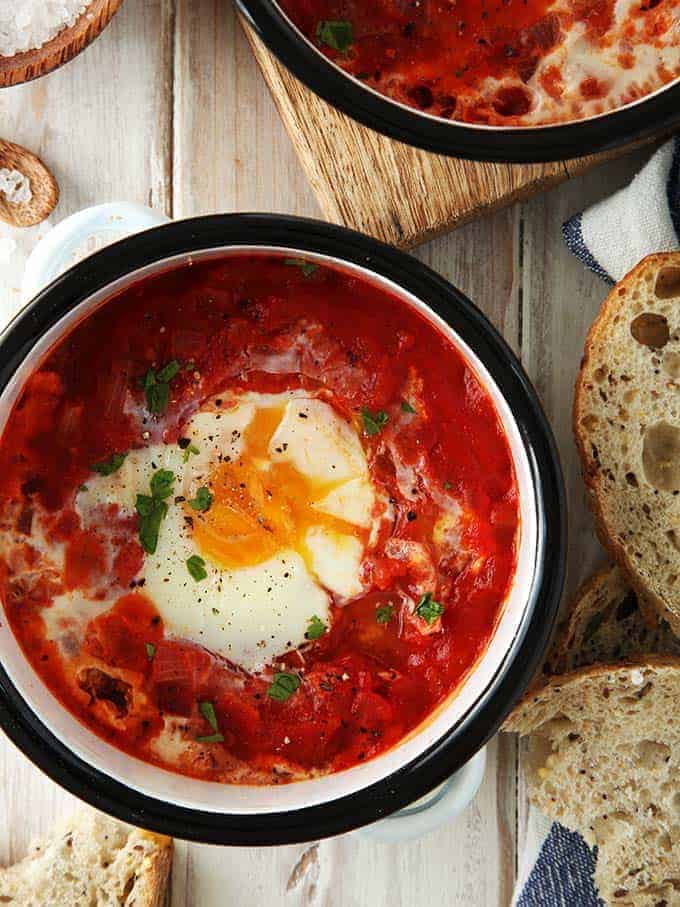 Egg baked in tomato sauce in a round bowl shot from above with slices of rustic bread at borders.