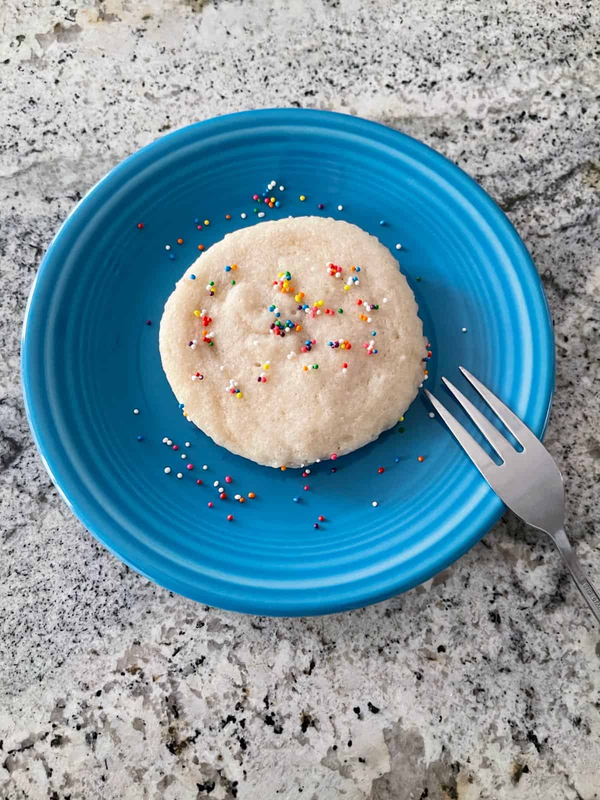 Vanilla mug cake topped with rainbow sprinkles on blue plate with fork.