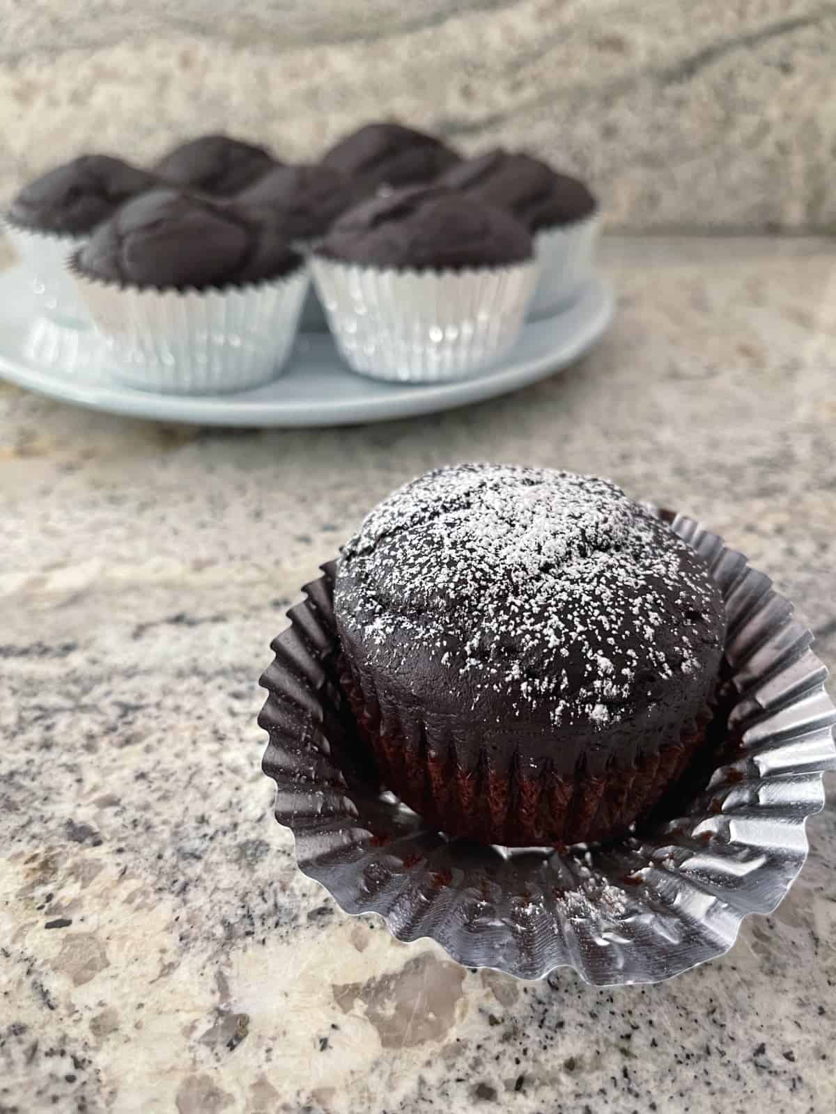 Chocolate cupcake dusted with powdered sugar in front of plate of plain chocolate cupcakes.