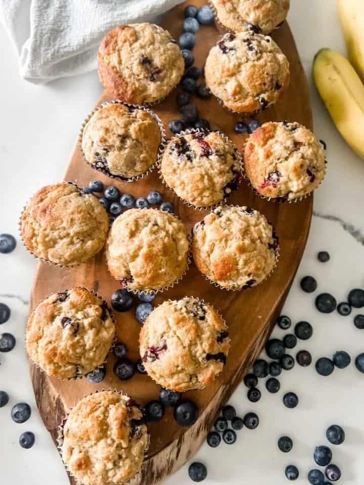 Blueberry Muffins on oval board with blueberries scattered around and bananas in background.