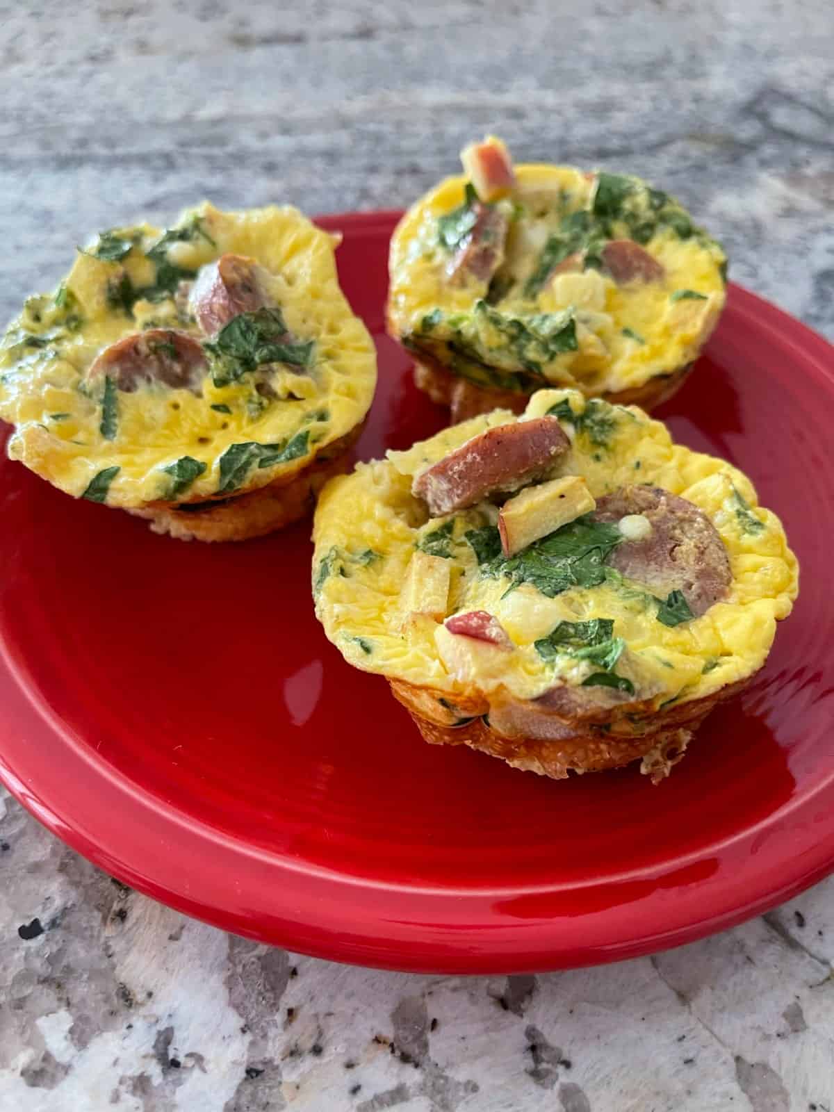 Three egg muffins with spinach and apple chicken sausage on small red plate on granite.