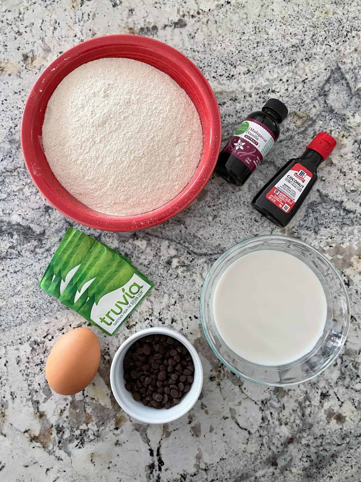 Ingredients including Kodiak Cakes mix, Truvia packets, egg, mini chocolate chips, almond milk, vanilla and coconut extract on granite.