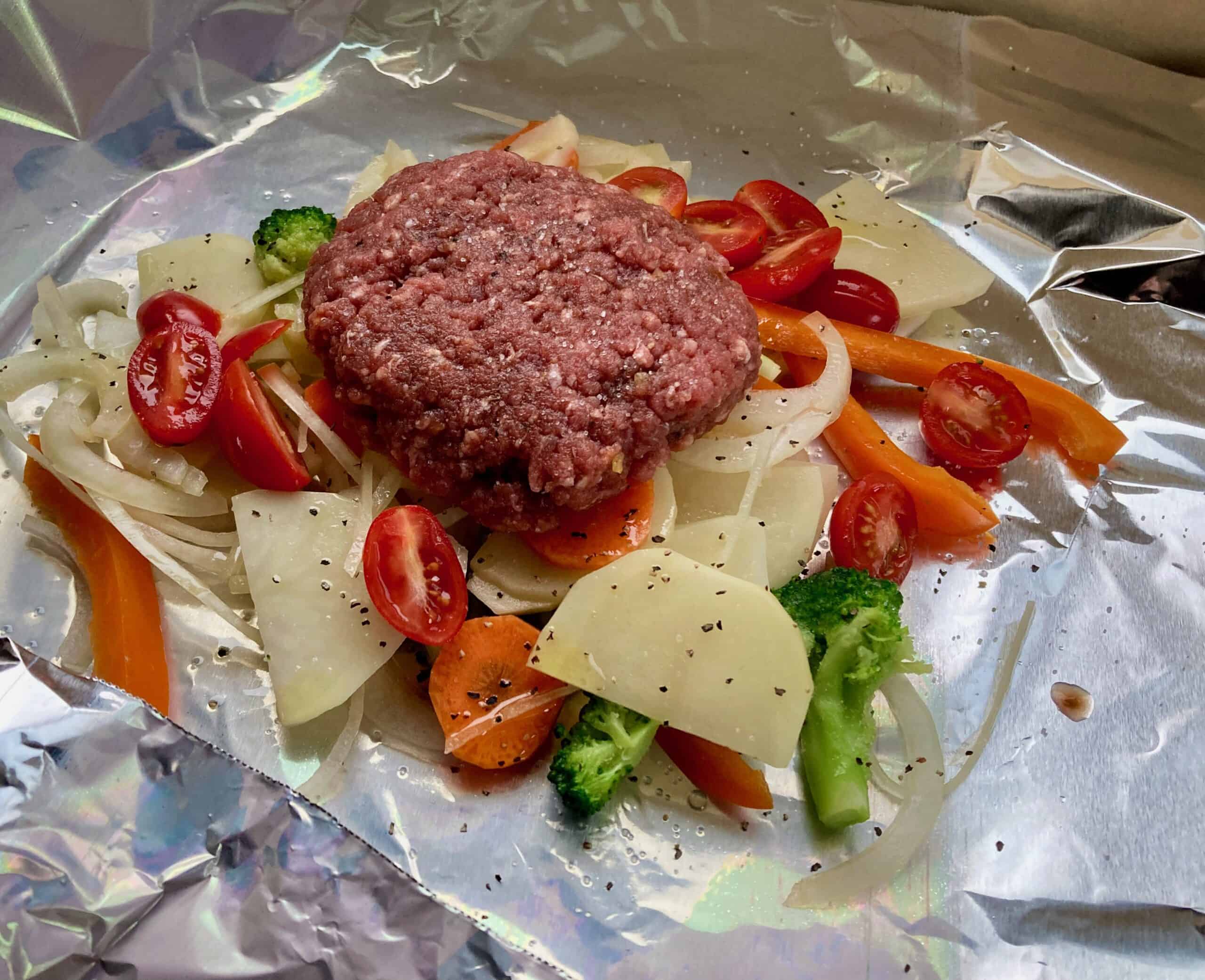 Assorted vegetables and ground beef patty on a sheet of heavy duty aluminum foil.