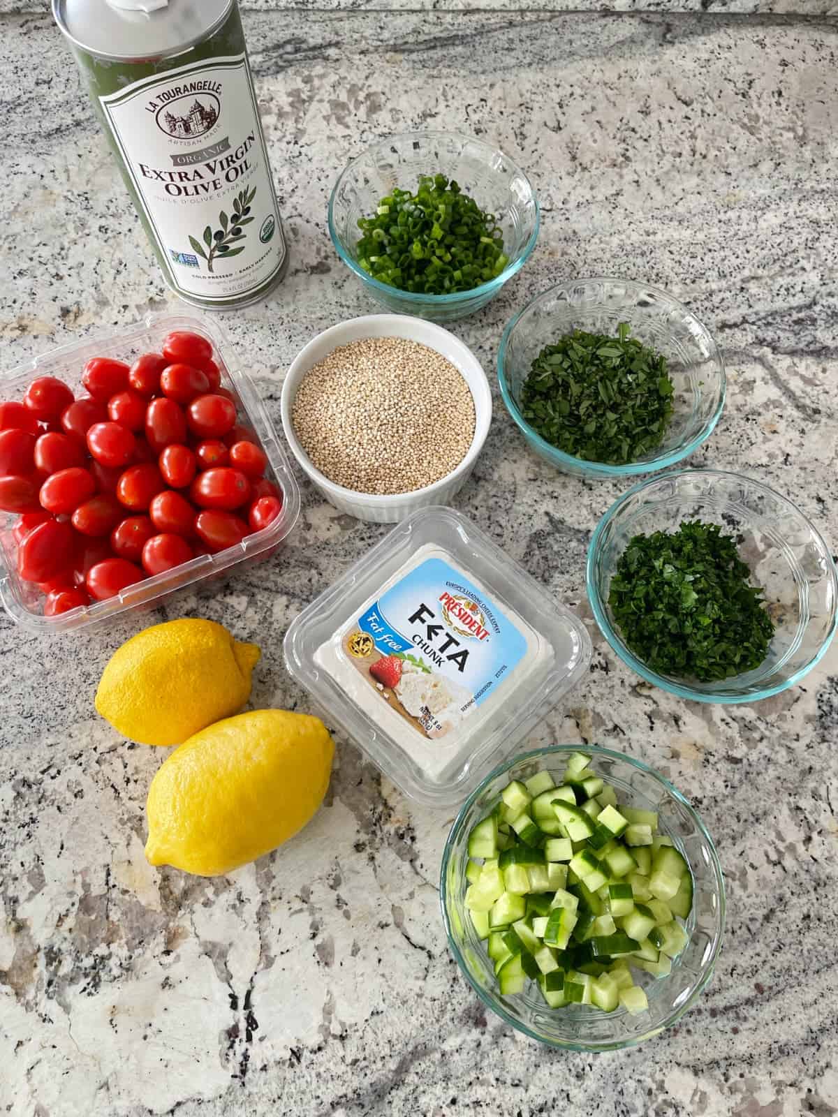 Ingredients including chopped cucumber, two lemons, cherry tomatoes, fat free feta cheese, quinoa, green onions, parsley, mint and bottle of olive oil on granite counter.