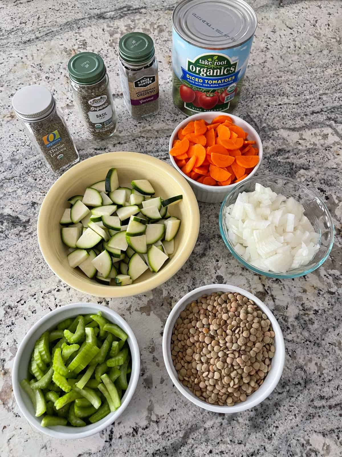 Ingredients including dry green lentils, chopped celery, carrots, zucchini and onions, canned diced tomatoes, dried basil, dried thyme and dried oregano on granite.