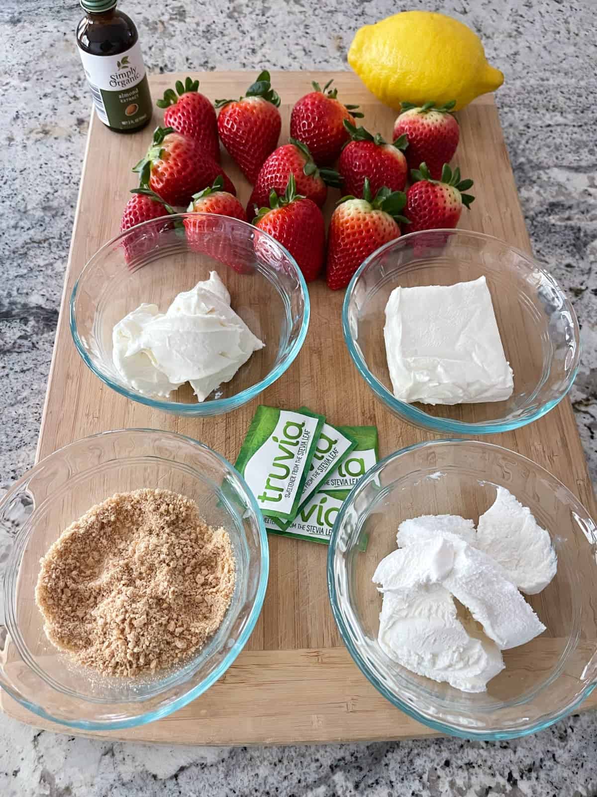Fresh strawberries, almond extract, whole lemon, Truvia sweetener packets, graham cracker crumbs, light cream cheese and lightly whipped topping on a wooden cutting board.