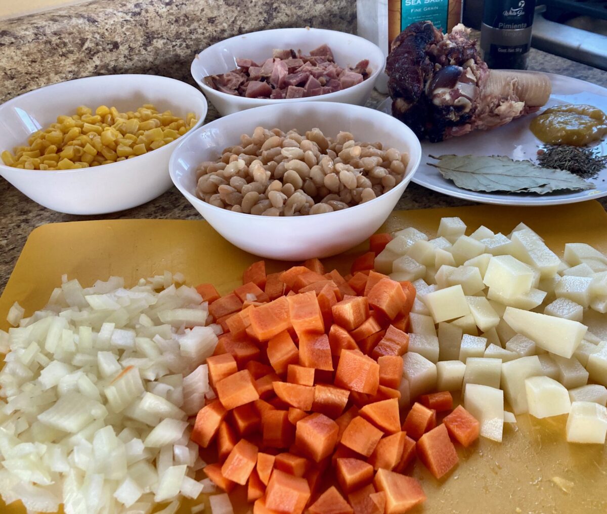 Yellow Cutting Board with chopped onion, carrot, potato, bowls of white beans, corn, chopped ham, ham bone, spices