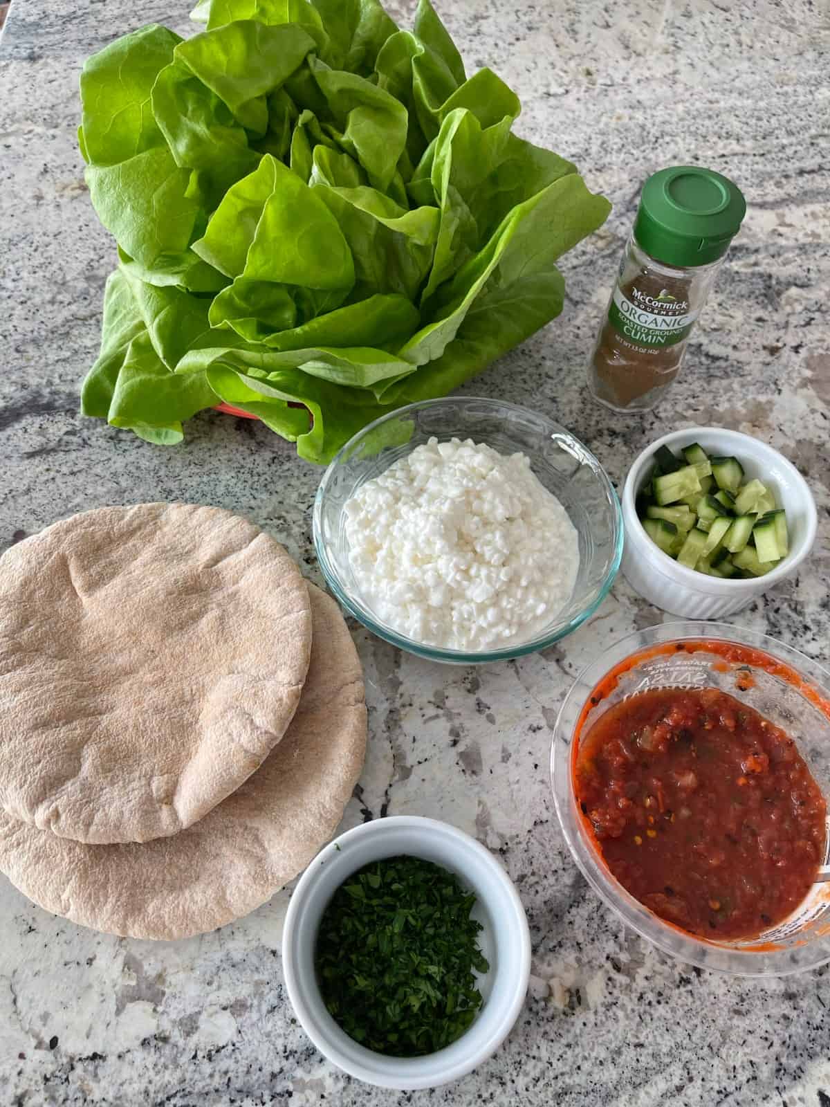 Bibb lettuce, cottage cheese, salsa, chopped cilantro leaves, two whole wheat pita breads, cucumber and ground cumin on a granite counter top.