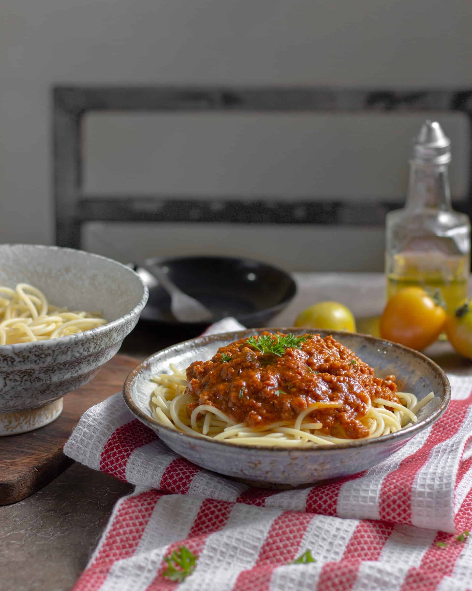 Bowl of spaghetti bolognese on red and white striped cloth with olive oil and plain pasta bowl in the background.