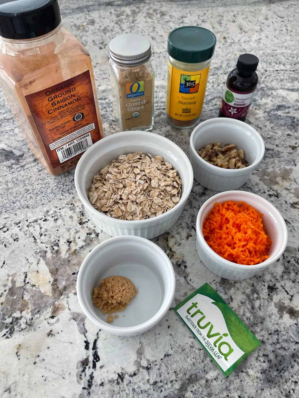 Rolled oats, shredded carrots, chopped walnuts, Truvia packet, brown sugar, ground cinnamon, ground ginger, ground nutmeg and vanilla extract on granite counter.