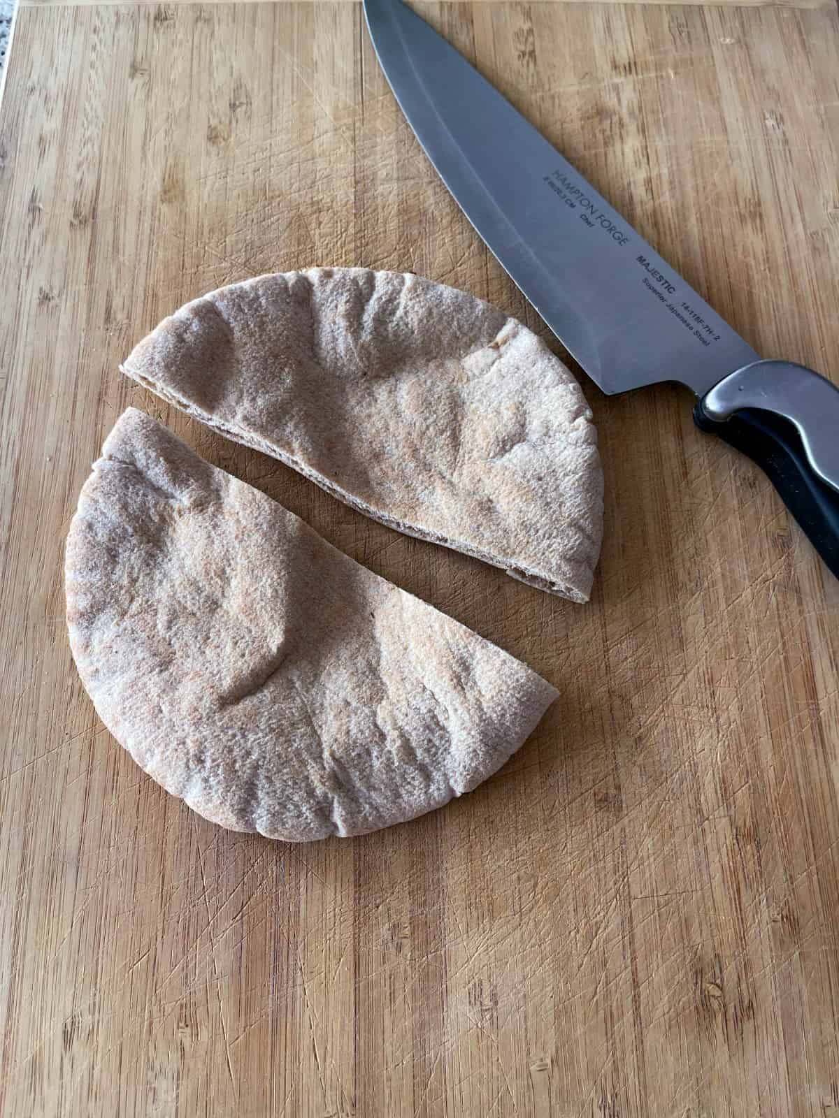 A whole wheat pita cut in half with a knife on a bamboo board.