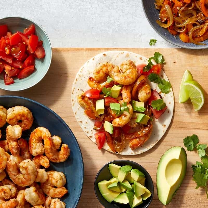wooden board with shallow bowl of shrimp, bowl of tomatoes, bowl of avocado and open shrimp fajita arranged on tortilla 