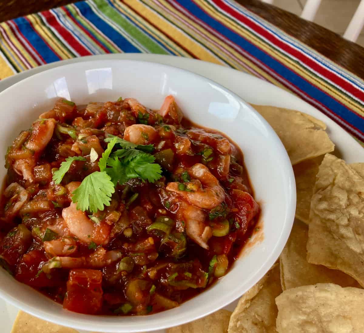 Shrimp Salsa Garnished with Cilantro White Bowl Surrounded by Tortilla Chips on Colorful Striped Mat.