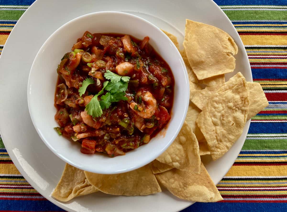 Shrimp salsa in a white bowl on a white plate with corn tortilla chips from above.