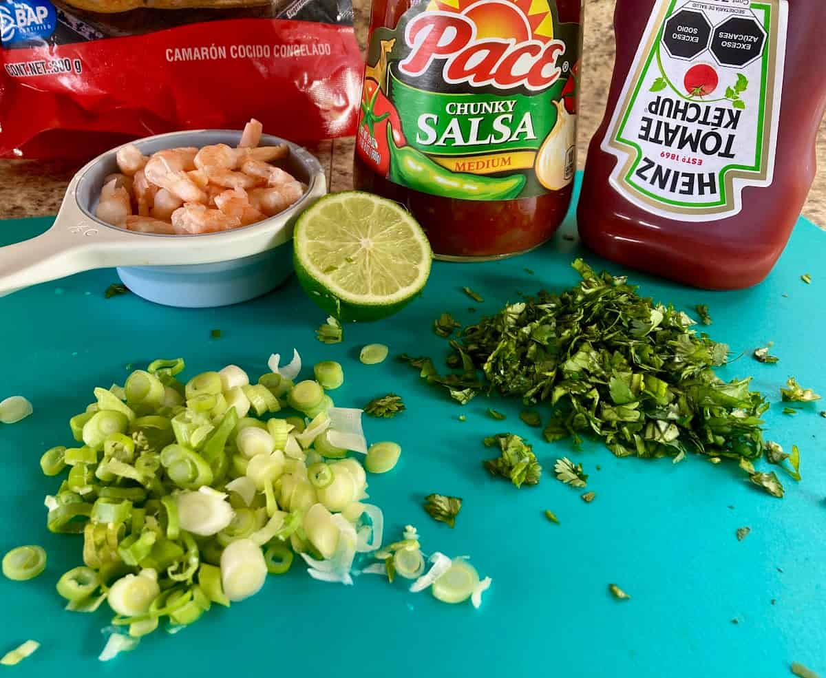 Chopped green onion, chopped cilantro, ½ lime on green cutting board with ½ cup small shrimp, prepared salsa and ketchup behind.