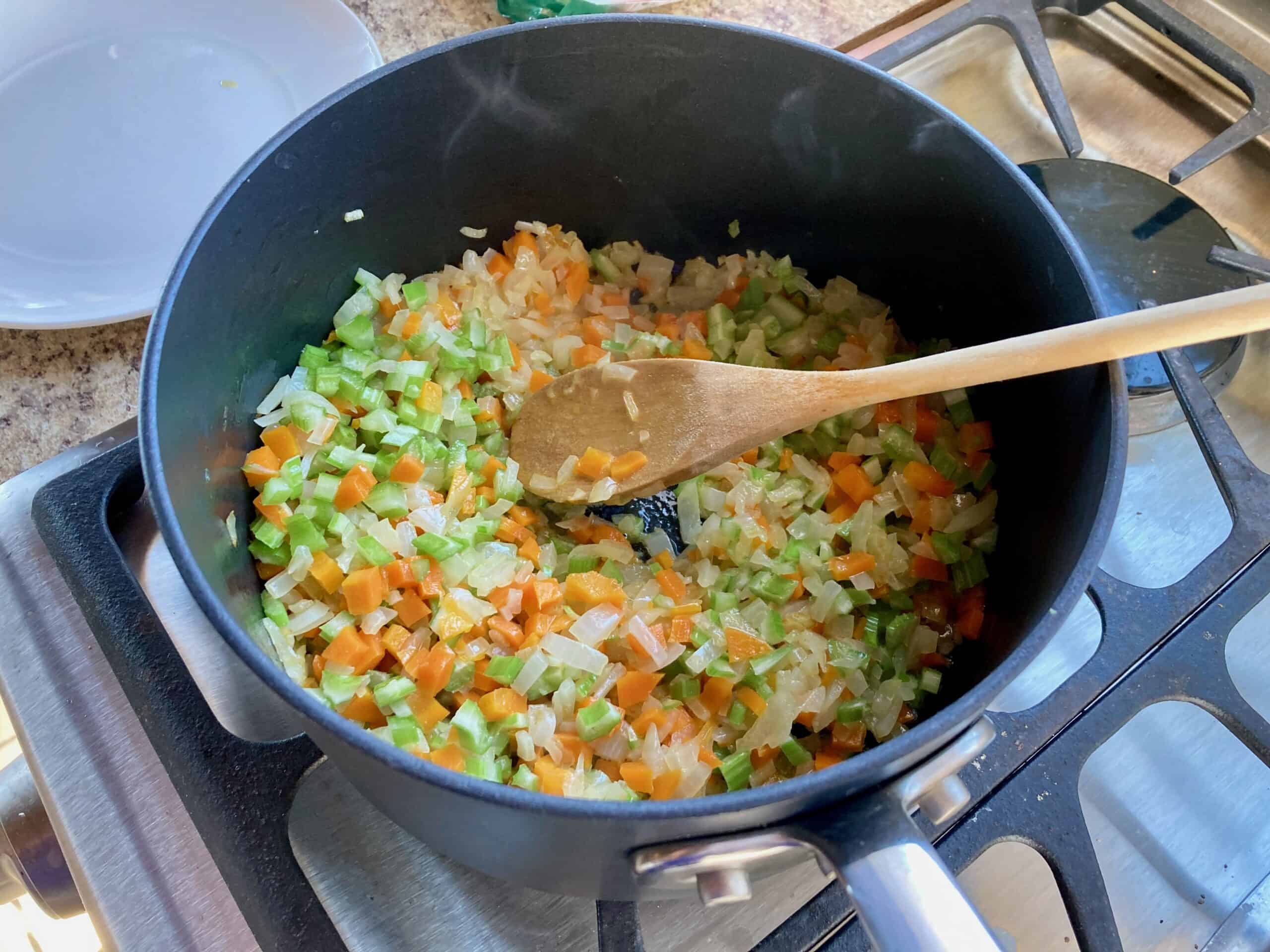 Chopped carrots, celery, onion and garlic in a black pot on the stove with a wooden spoon