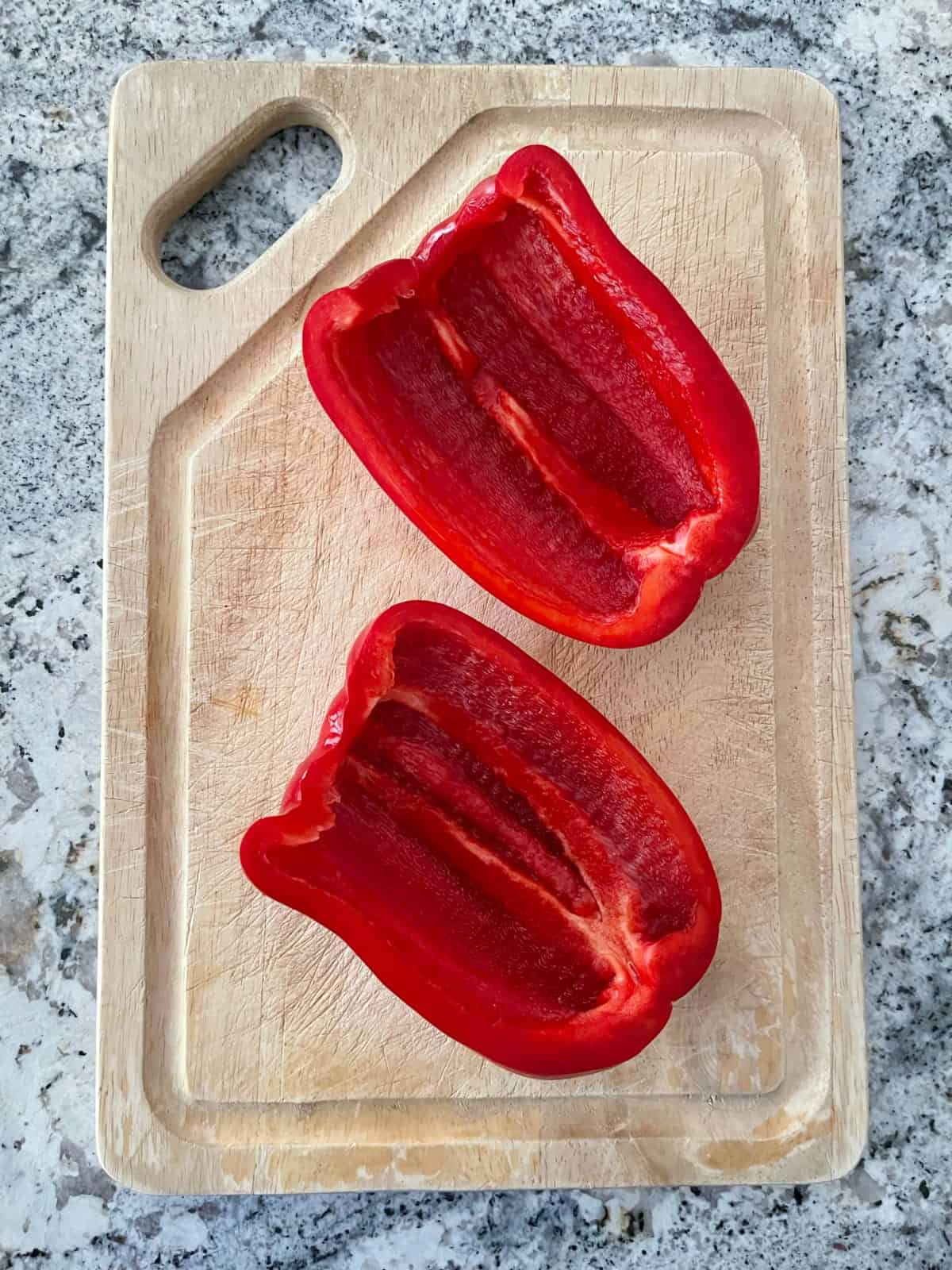 One red bell pepper halved lengthwise with stem, seeds and white membranes removed on wood cutting board.