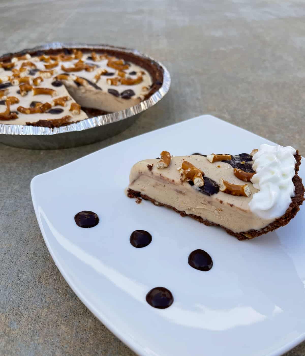 Slice of peanut butter pretzel pie with whipped topping on small white plate in front of whole pie.