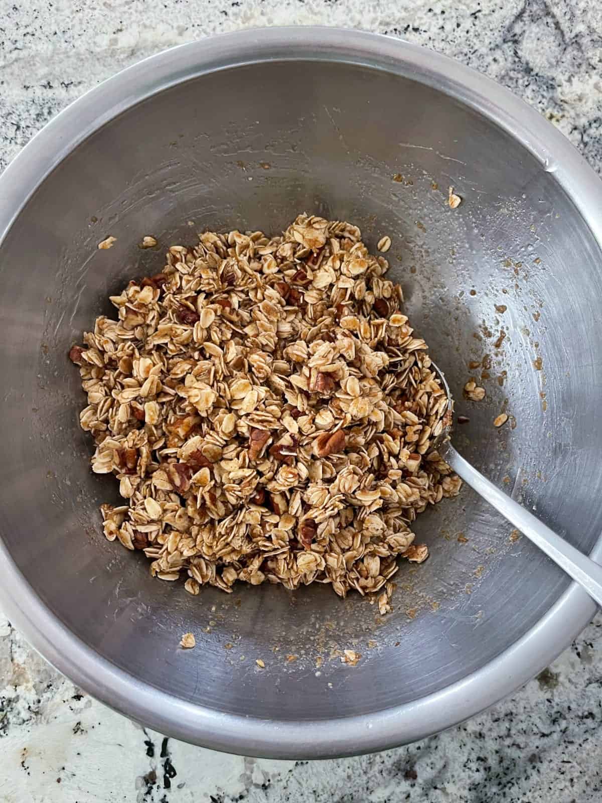 Mixing granola ingredients, including rolled oats, chopped pecans, flax meal, cinnamon, apple juice, brown rice syrup, oil and vanilla in mixing bowl with spoon.