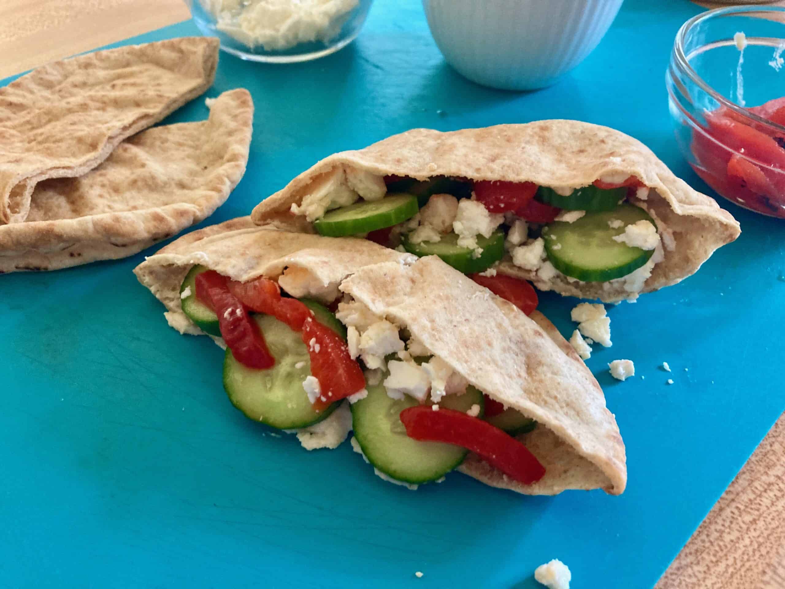 Two veggie hummus stuffed pitas on teal cutting board with small bowls of hummus and feta cheese in the background.