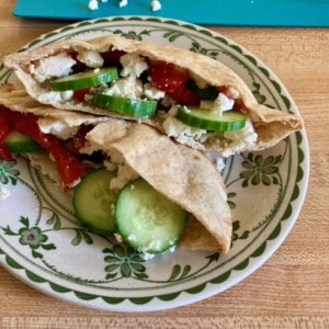 Two pita bread halves stuffed with hummus, slicked cucumber, roasted red bell pepper and feta cheese on flower rimmed plate