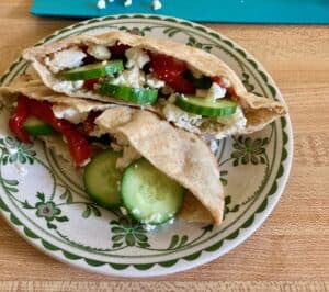 Two pita bread halves stuffed with hummus, slicked cucumber, roasted red bell pepper and feta cheese on flower rimmed plate