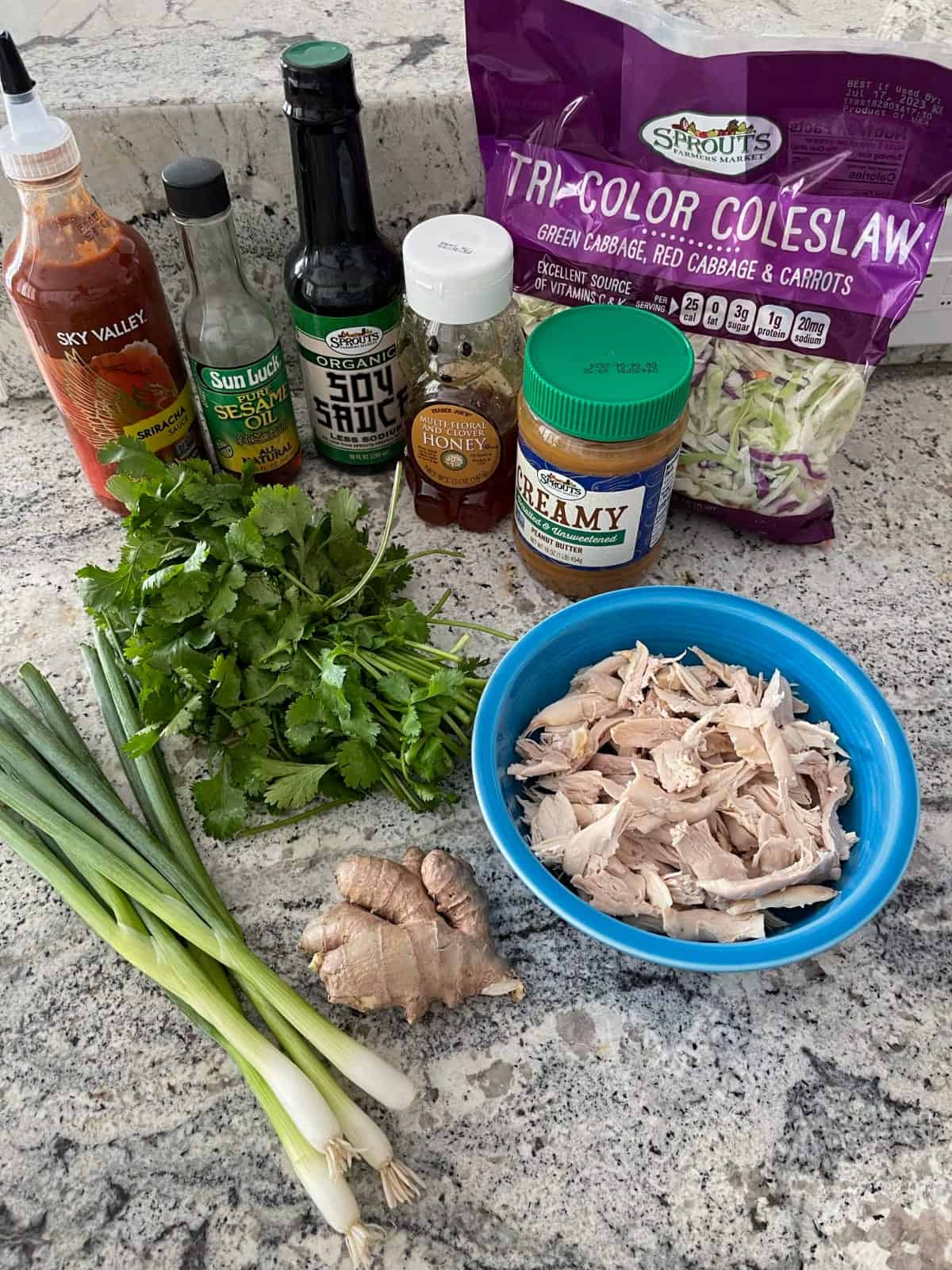 Ingredients including mixed tricolor slaw, shredded chicken breast, ginger root, green onion, cilantro, peanut butter, honey, low sodium soy sauce, sesame oil and Sriracha chili sauce on a granite counter.