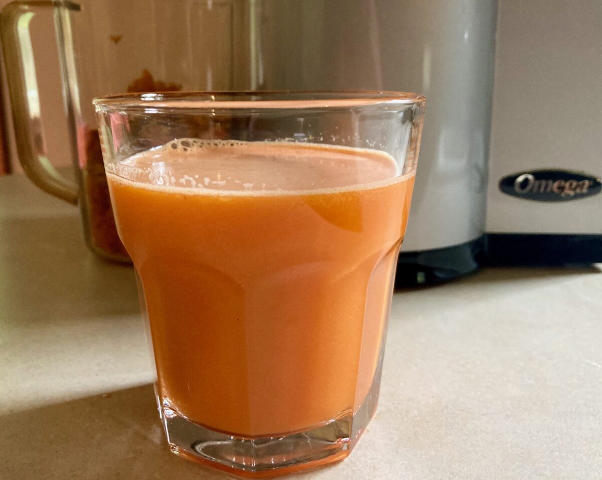 Glass of carrot beet pineapple juice in front of vertical Omega Juicer on counter.