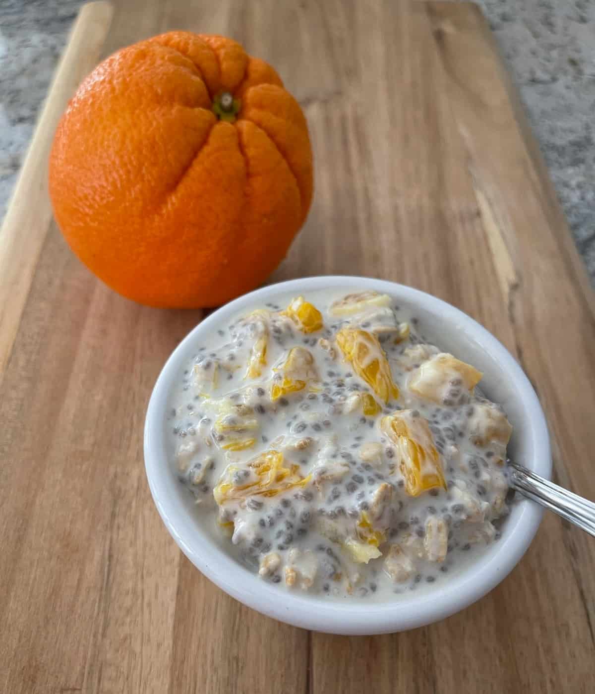 Overnight chia orange oats in small white bowl with spoon and fresh whole orange in the background on wood cutting board.