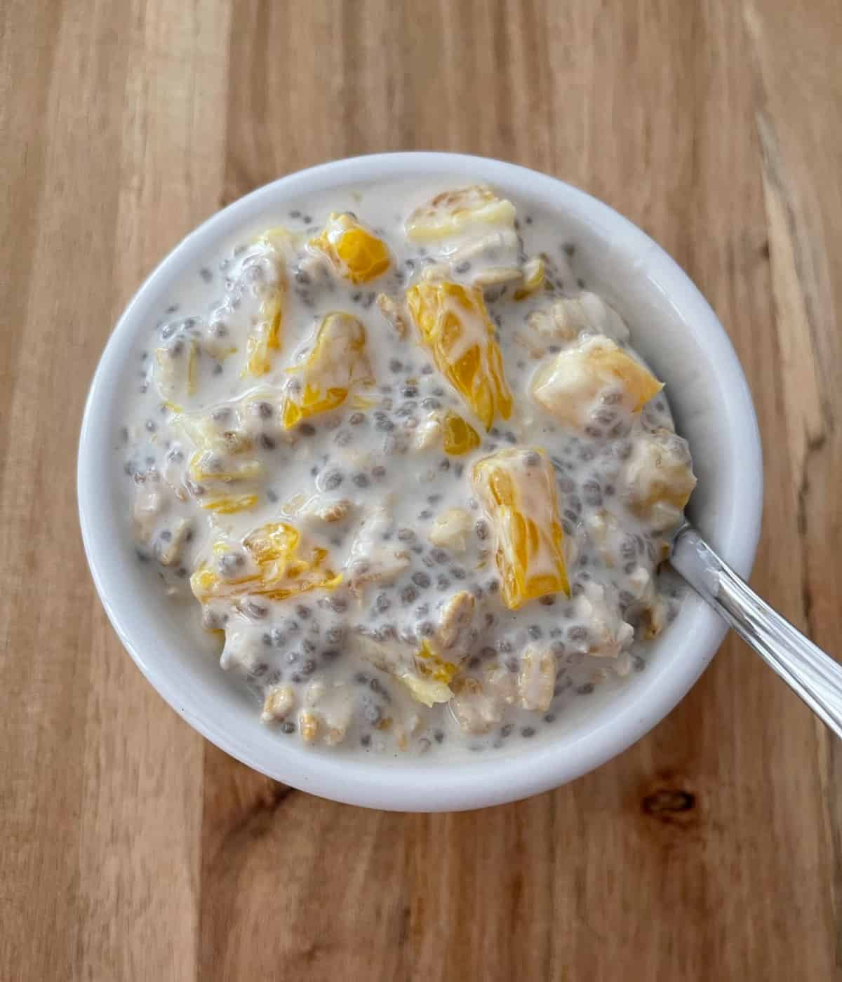Overnight orange oats with chia seeds in small white bowl with spoon on wood surface.