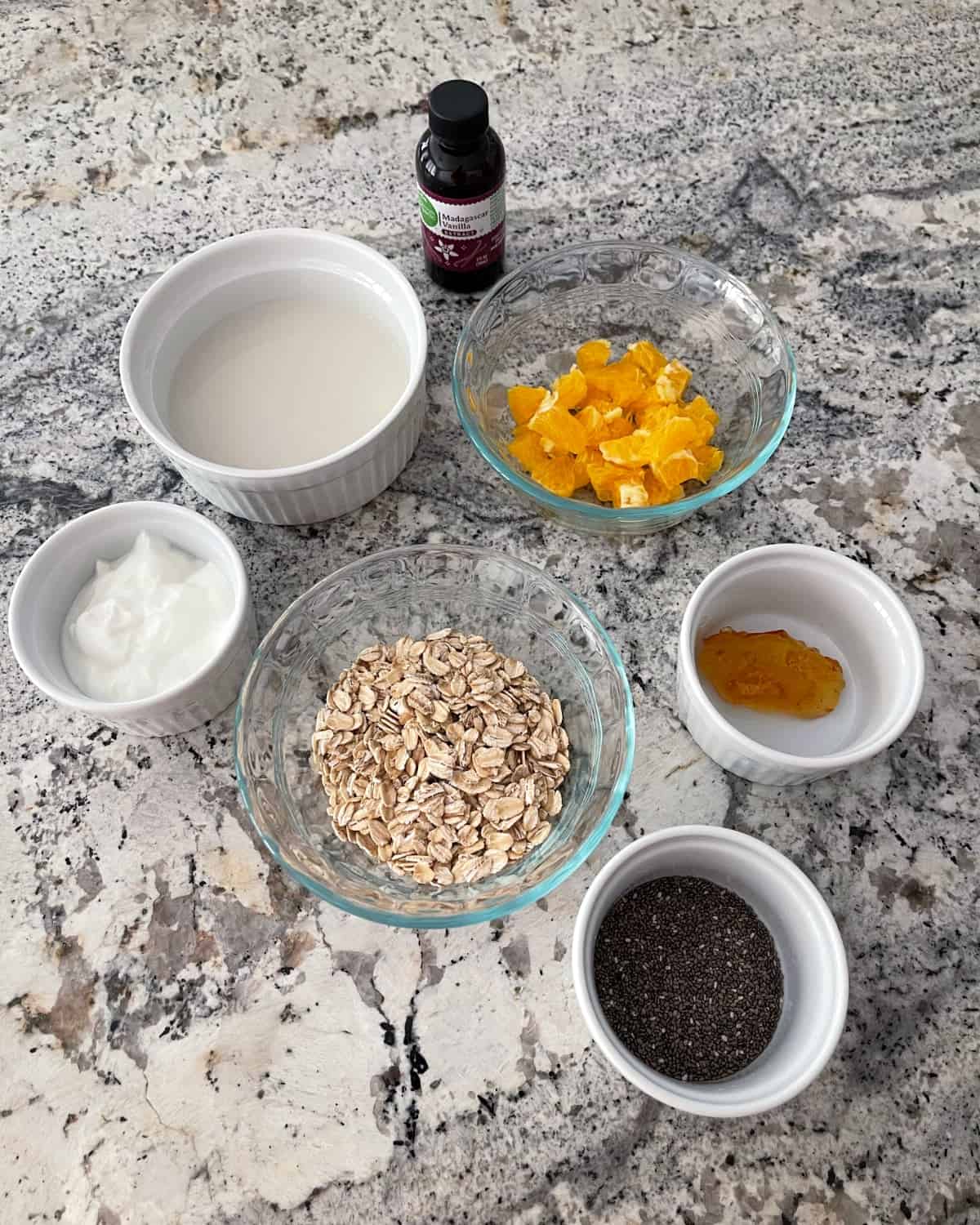 Ingredients including plain nonfat Greek yogurt, almond milk, vanilla extract, chopped orange, rolled oats, orange marmalade and chia seeds on granite counter.