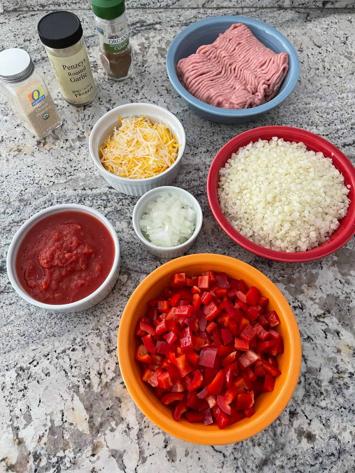 Ingredients including chopped red bell peppers, riced cauliflower, ground turkey, crushed tomatoes, chopped onion, shredded cheese and spices on granite.