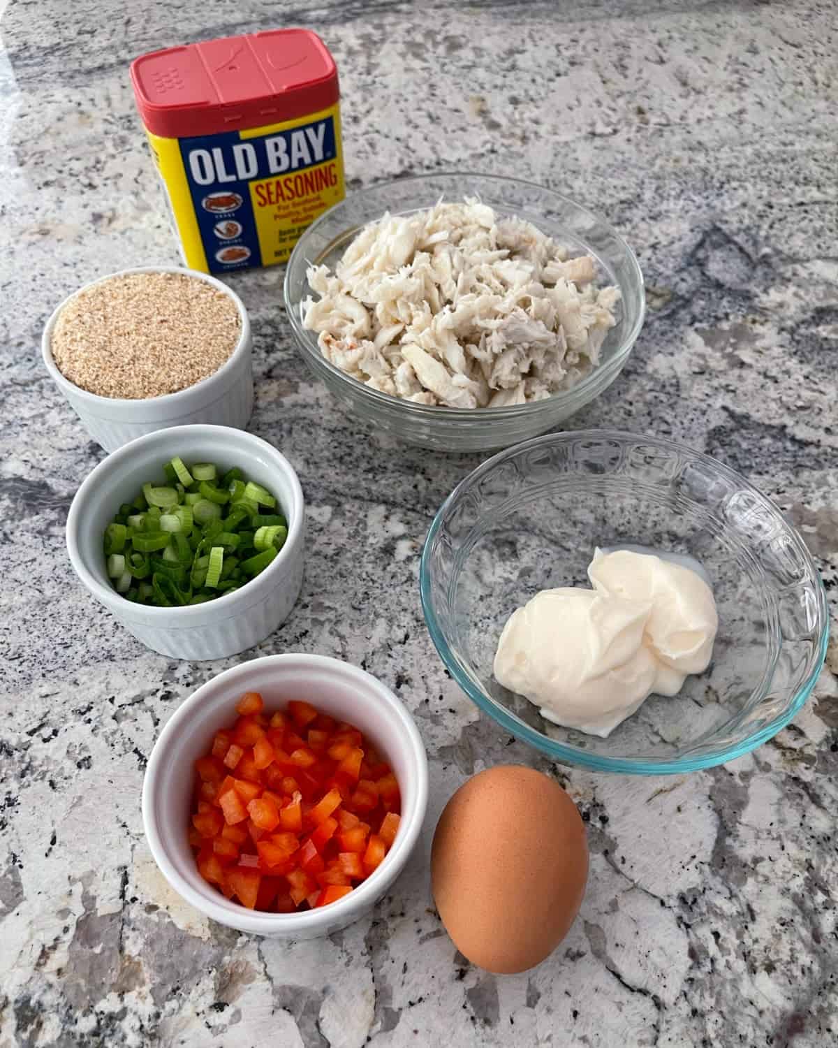 Ingredients including Old Bay seasoning, lump crabmeat, light mayonnaise, egg, diced red bell pepper, sliced green onion and breadcrumbs.