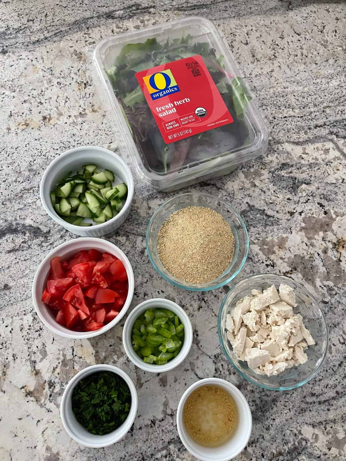Ingredients included package of mixed salad greens, chopped cucumber, chopped tomato, chopped celery, chopped Italian parsley, minced garlic, crumbled tofu and dry falafel mix on granite.