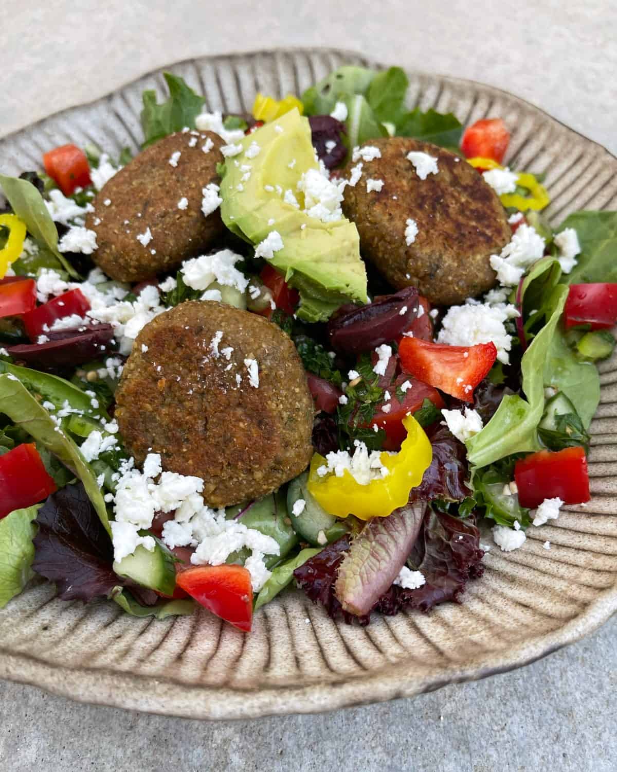 Tofu-falafel salad on bed of mixed greens with sliced avocado, Kalamata olives, chopped red bell pepper, yellow banana pepper rings and crumbled feta cheese.