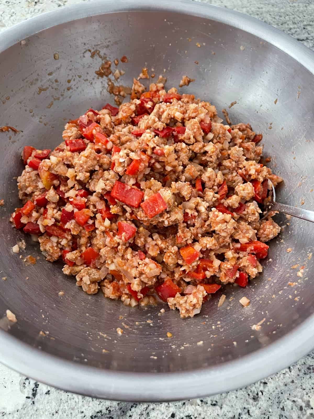 Mixing unstuffed pepper casserole ingredients - cooked ground turkey, chopped bell peppers, riced cauliflower and spices in mixing bowl with spoon.