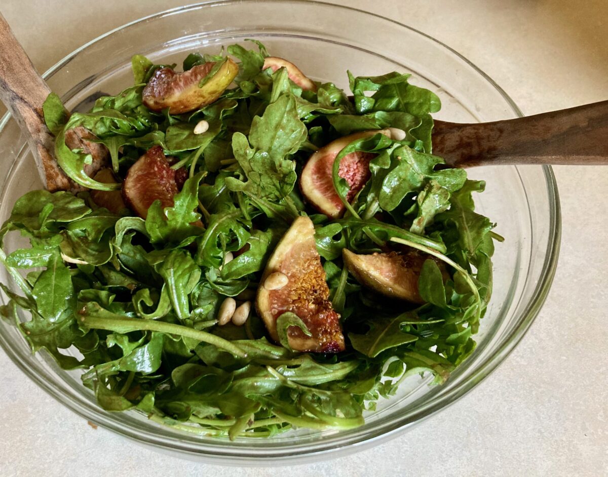 Arugula Fig Salad in a large clear glass bowl with wooden salad spoons.